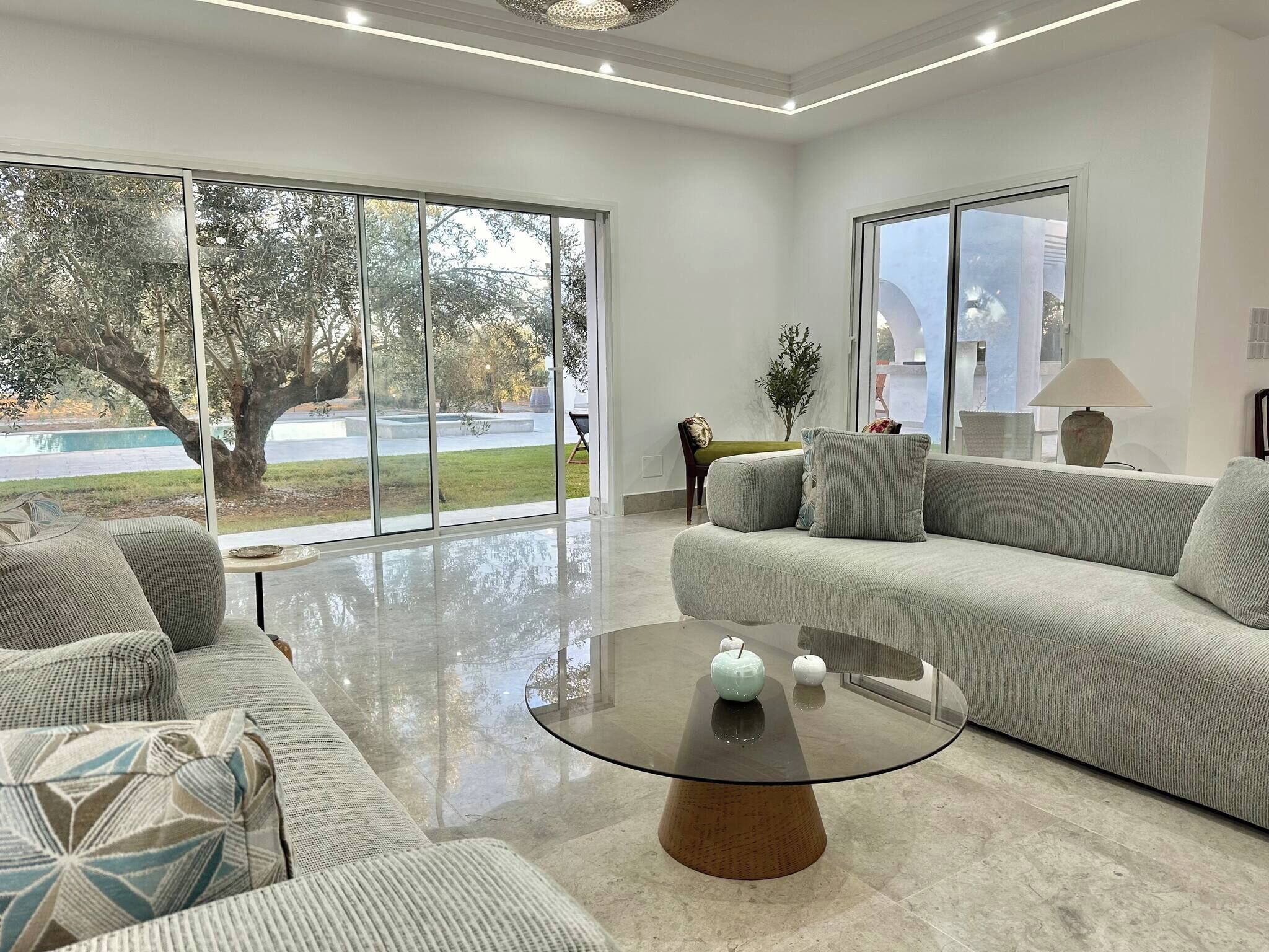 A modern living room with a large sofa, a glass coffee table, and a view of a tree through large windows.