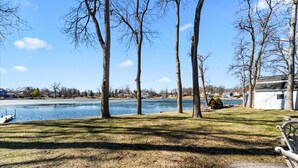 A lakeside view with trees, a dock, and a house in the background. - Lakefront Ranch | Hot Tub · 30-Day Min Stay (Waterford Township)