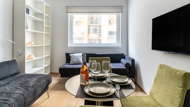 Ein modernes Wohnzimmer mit Esstisch für vier Personen, einer Couch, einem Bücherregal und einem großen Fenster mit Blick auf die Stadt.