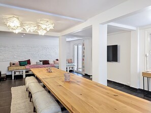 A modern living room with a wooden dining table, benches, a white brick wall, and a hanging light fixture. - HENRI CAMILLE RE-  Beach House - 2 bedrooms house - Garden - 3 min beaches (Cannes)