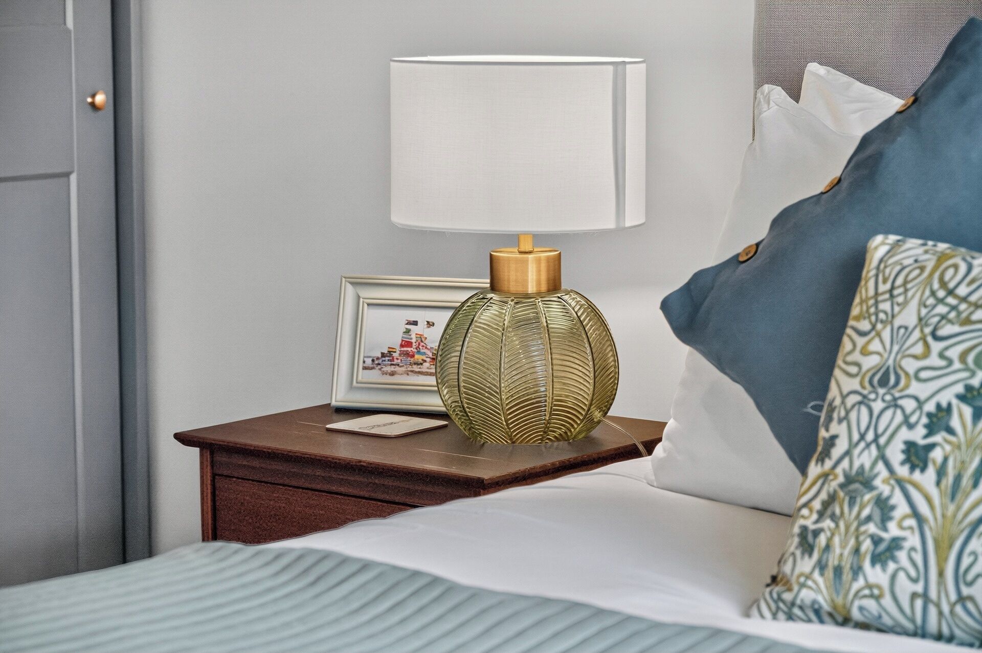 A neatly made bed with a white pillow and a patterned pillow, a bedside table with a lamp and a framed picture, and a door to the left.