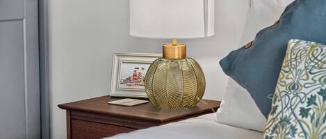 A neatly made bed with a white pillow and a patterned pillow, a bedside table with a lamp and a framed picture, and a door to the left.