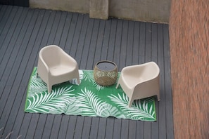 Un petit espace extérieur aménagé avec deux chaises blanches et une table en osier, le tout posé sur un tapis à motifs de feuilles vertes.
