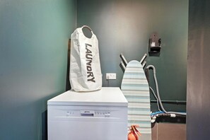 Ein Waschbereich mit Waschmaschine, Bügelbrett und einer mit 'Wäsche' beschrifteten Tasche.