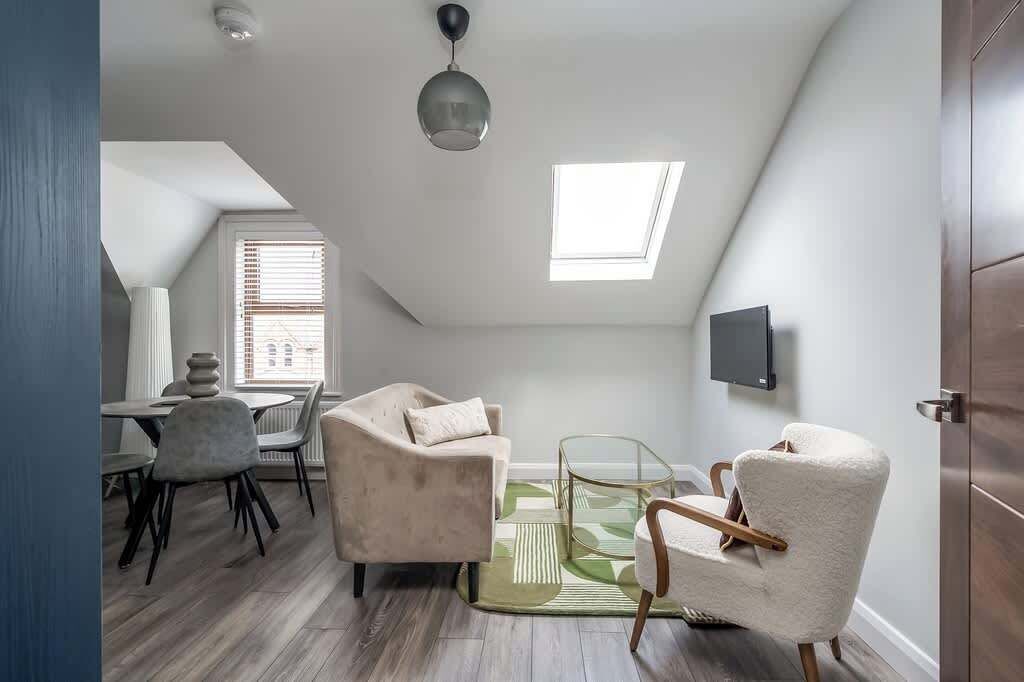 A loft-style room with a dining area, two armchairs, a glass coffee table, a ceiling fan, a TV mounted on the wall, and skylights.