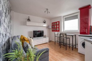Un salon moderne avec un canapé gris, un téléviseur à écran plat, un meuble blanc, un mur décoré de tableaux encadrés de rouge et un lave-linge.