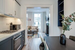 A modern kitchen with a gas stove, microwave, and cabinets. There is a dining area with a table and chairs, and a sofa in the background. - Central Belfast Apartments Queens Lofts (Belfast)