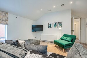 Un salon moderne avec un téléviseur à écran plat, un canapé vert, des fauteuils gris et un tapis à motifs.