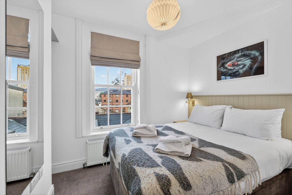 Ein Hotelzimmer mit Bett, Fenster mit Blick auf Gebäude, einem Bild an der Wand und einer Deckenleuchte.