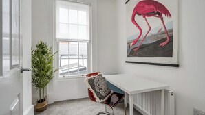 Ett rum med ett vitt skrivbord, en röd stol med ett mönstrat kuddöverdrag, ett stort fönster, en krukväxt och ett väggmonterat konstverk som föreställer en flamingo.
