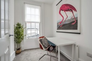 Ein Zimmer mit einem weißen Schreibtisch, einem roten Stuhl mit gemustertem Sitzkissen, einem großen Fenster, einer Topfpflanze und einem Wandbild einer Flamingo.