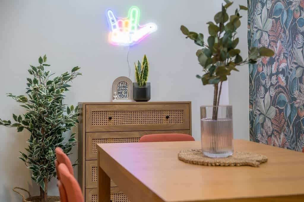A dining area with a wooden table, a neon sign, and two potted plants.