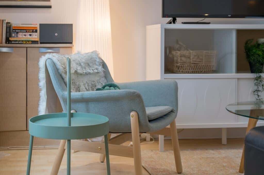 A modern living room with a light blue armchair, a small round table, a bookshelf with books and a laptop, a TV on a white cabinet, and a plant on the right.