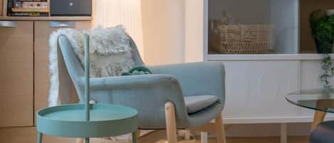 A modern living room with a light blue armchair, a small round table, a bookshelf with books and a laptop, a TV on a white cabinet, and a plant on the right.