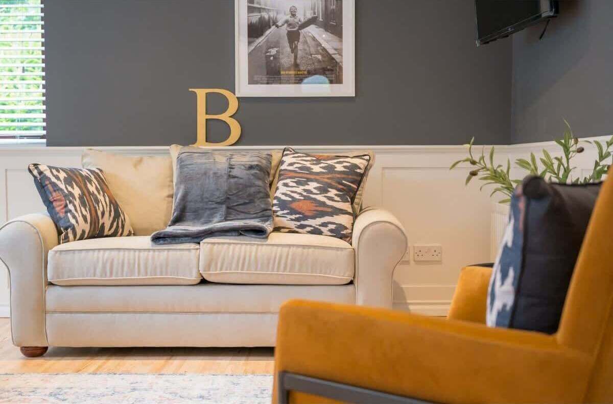 Een woonkamer met een beige bank, gekleurde kussens, een gele fauteuil en een lijst met de letter 'B' aan de muur.