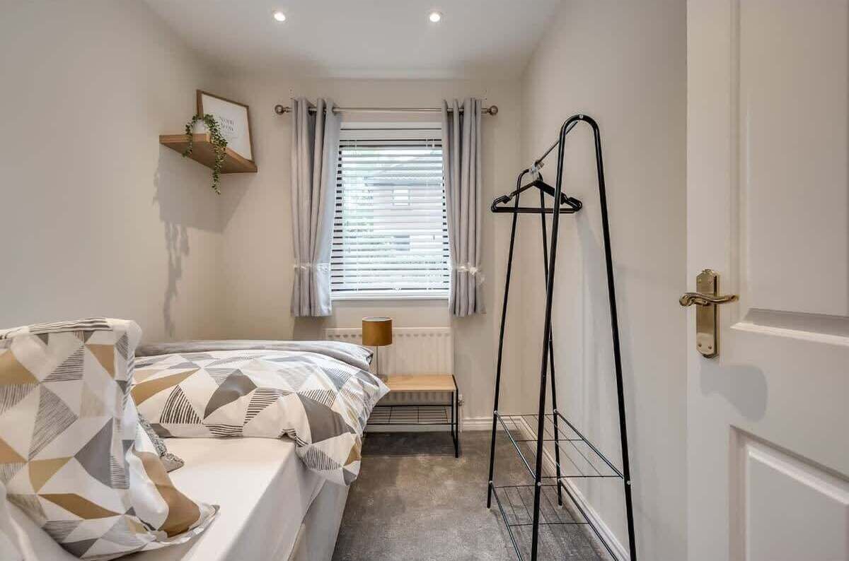 A bedroom with a bed, a radiator, a window with curtains, a wall-mounted shelf with a plant, and a door with a handle.