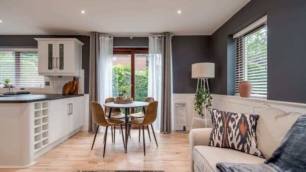 Een moderne keuken met witte kastjes, een eettafel met stoelen, een bank met gekleurde kussens en een vloerlamp.