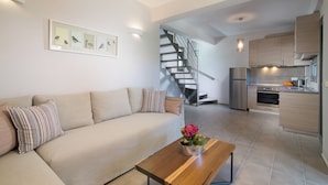 Una sala moderna con un sofá, una mesa de centro con flores, y una escalera que lleva a una zona de cocina con un refrigerador y estufa.