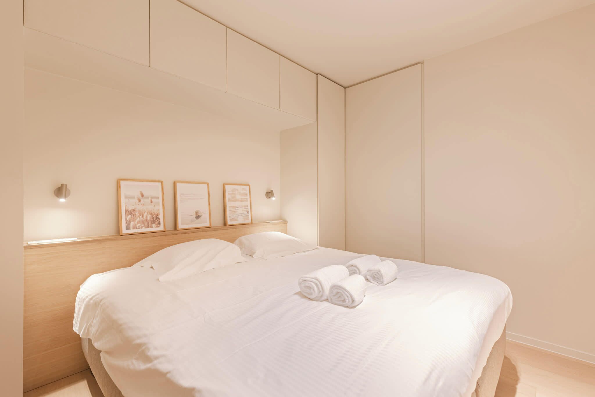 Un lit bien fait, avec du linge de lit blanc et des serviettes pliées, une tête de lit en bois et des tableaux encadrés accrochés au mur.