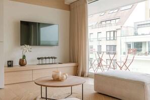 A modern living room with a flat-screen TV, a round coffee table with decorative items, a cushioned seating area, and a view of a building through large windows. - Stylish coastal apartment for 4 with sea view (Knokke-Heist)