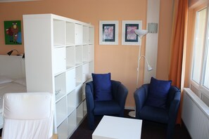 A room with a white bed, a white wardrobe, two blue armchairs, a white table, and two framed pictures on the wall.