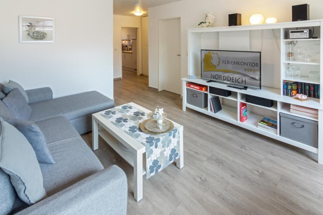Un salon avec un canapé gris, une table basse blanche ornée d’un motif floral, un téléviseur posé sur un meuble blanc et une bibliothèque contenant divers objets.