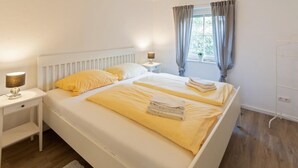 Une chambre à coucher avec un lit blanc, une table de chevet avec une lampe, une fenêtre avec des rideaux et un radiateur.