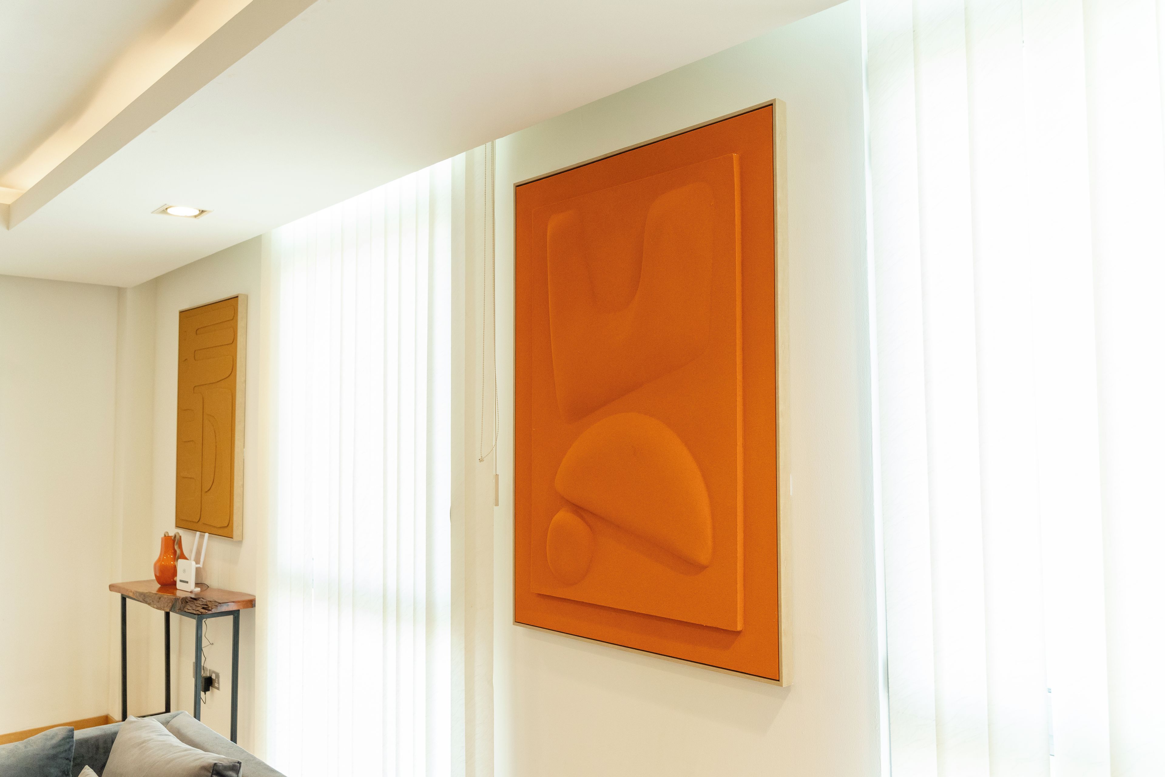 A room with a large orange artwork on the wall, a small table with a vase, and a sofa.