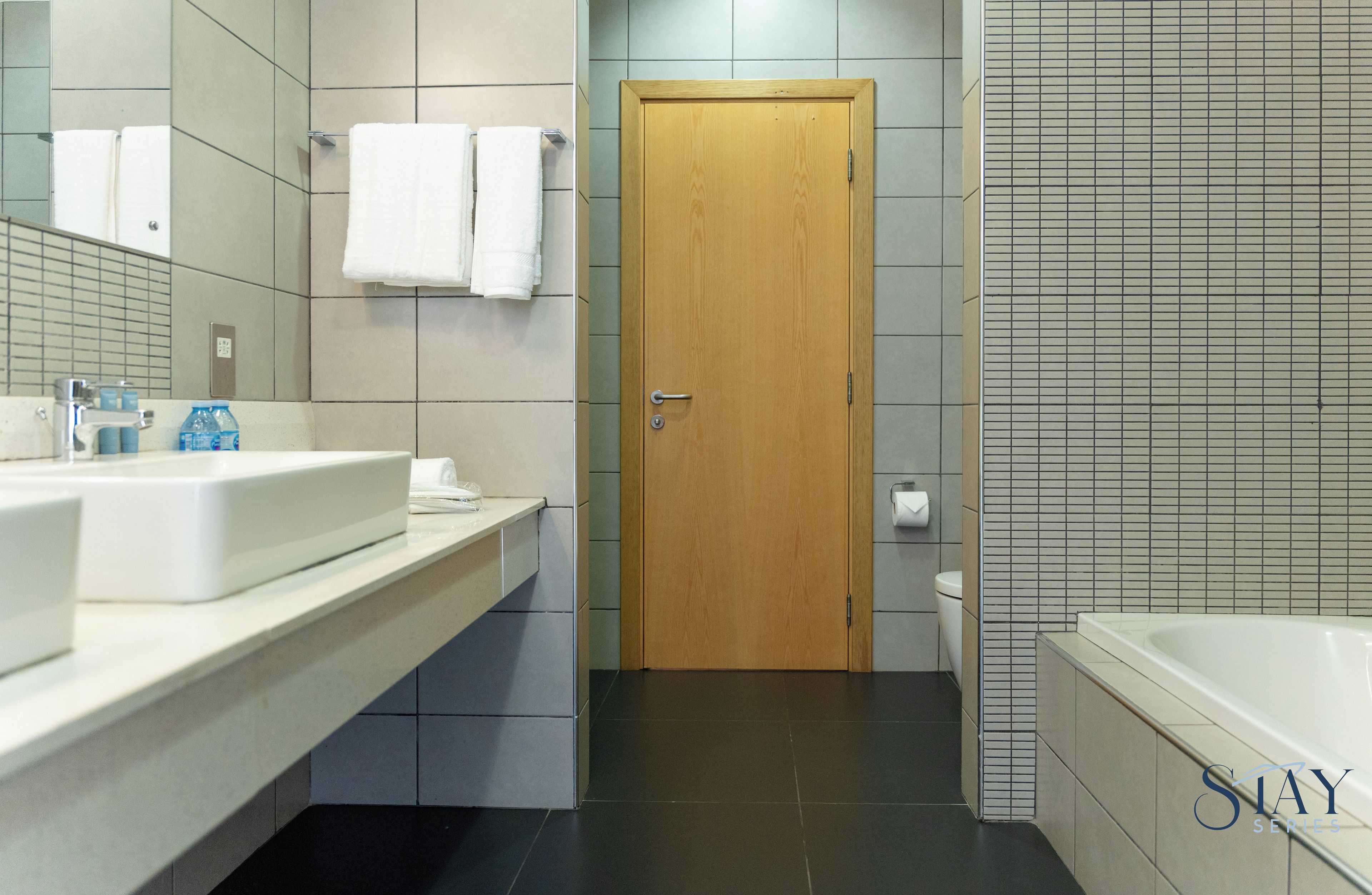A modern bathroom with a wooden door, a white sink, a bathtub, and a mirror.