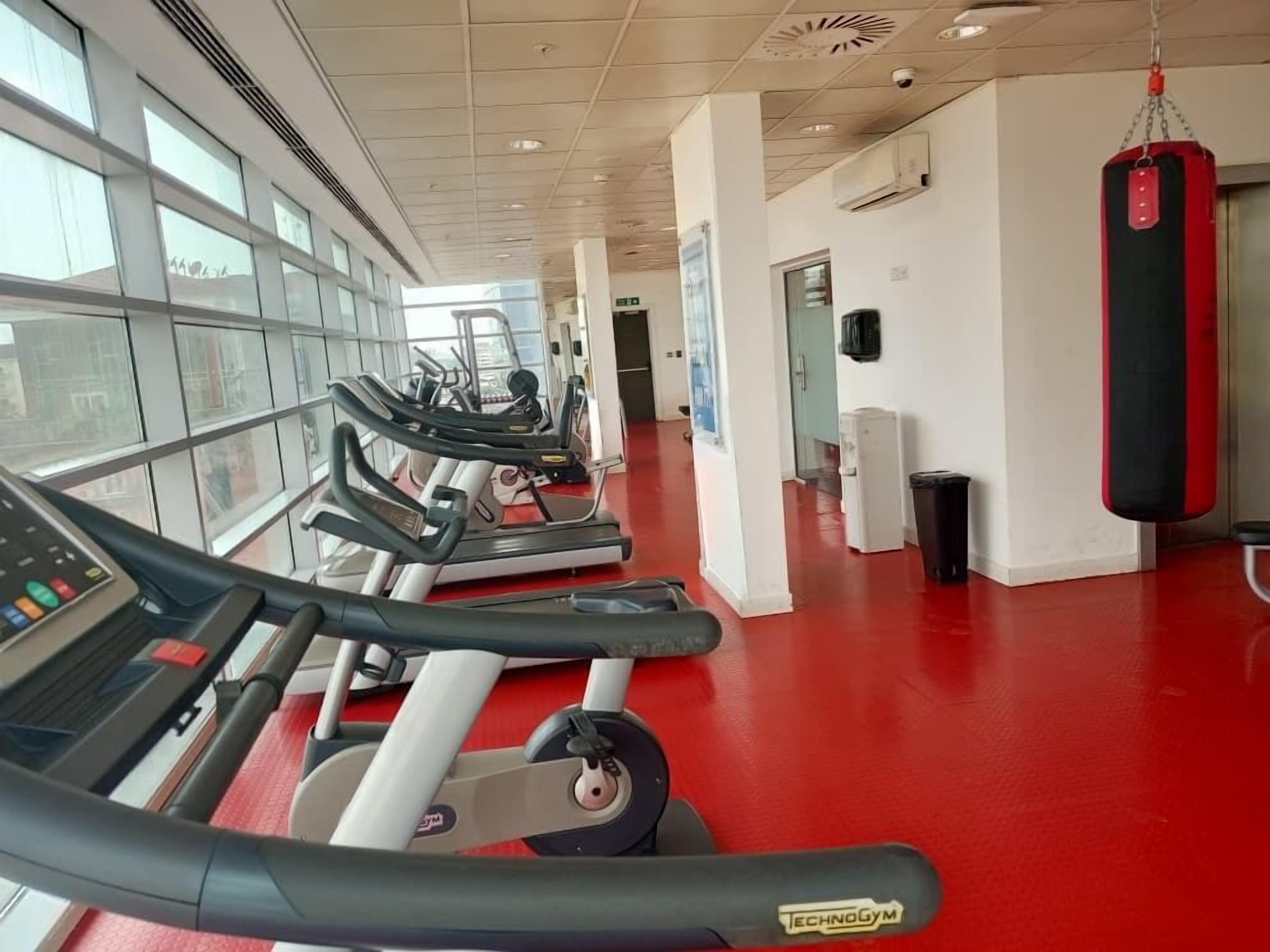 Un gimnasio con cintas de correr, un saco de boxeo y un muro con ventanas.