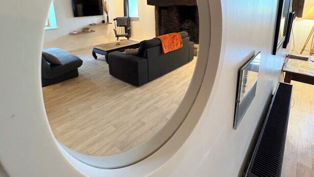 A circular window revealing a living room with a sofa, a fireplace, and a television. - Cosy and warm "l'Atelier des poutres" cottage (Guilliers)