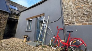 A building with a stone wall, a grey exterior, and a red bicycle parked outside. - Cosy and warm "l'Atelier des poutres" cottage (Guilliers)