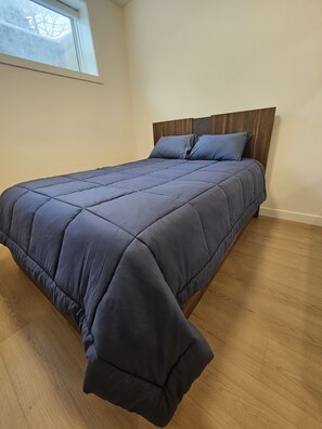 A bed with a wooden headboard, a dark blue quilt, and a window with a view of bare trees.