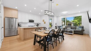 Eine moderne Küche mit einer zentralen Kochinsel, ein Essbereich mit Holztisch und Stühlen sowie ein Wohnbereich mit Sofa und Couchtisch.