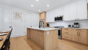Eine moderne Küche mit weißen Schränken, einer zentralen Kochinsel, Edelstahlgeräten und einem Essbereich mit einem Holztisch und Stühlen.