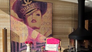 Un soggiorno moderno con un'ampia murale Chanel, una poltrona rosa, un tavolino in vetro con un vaso di fiori e un caminetto nero.
