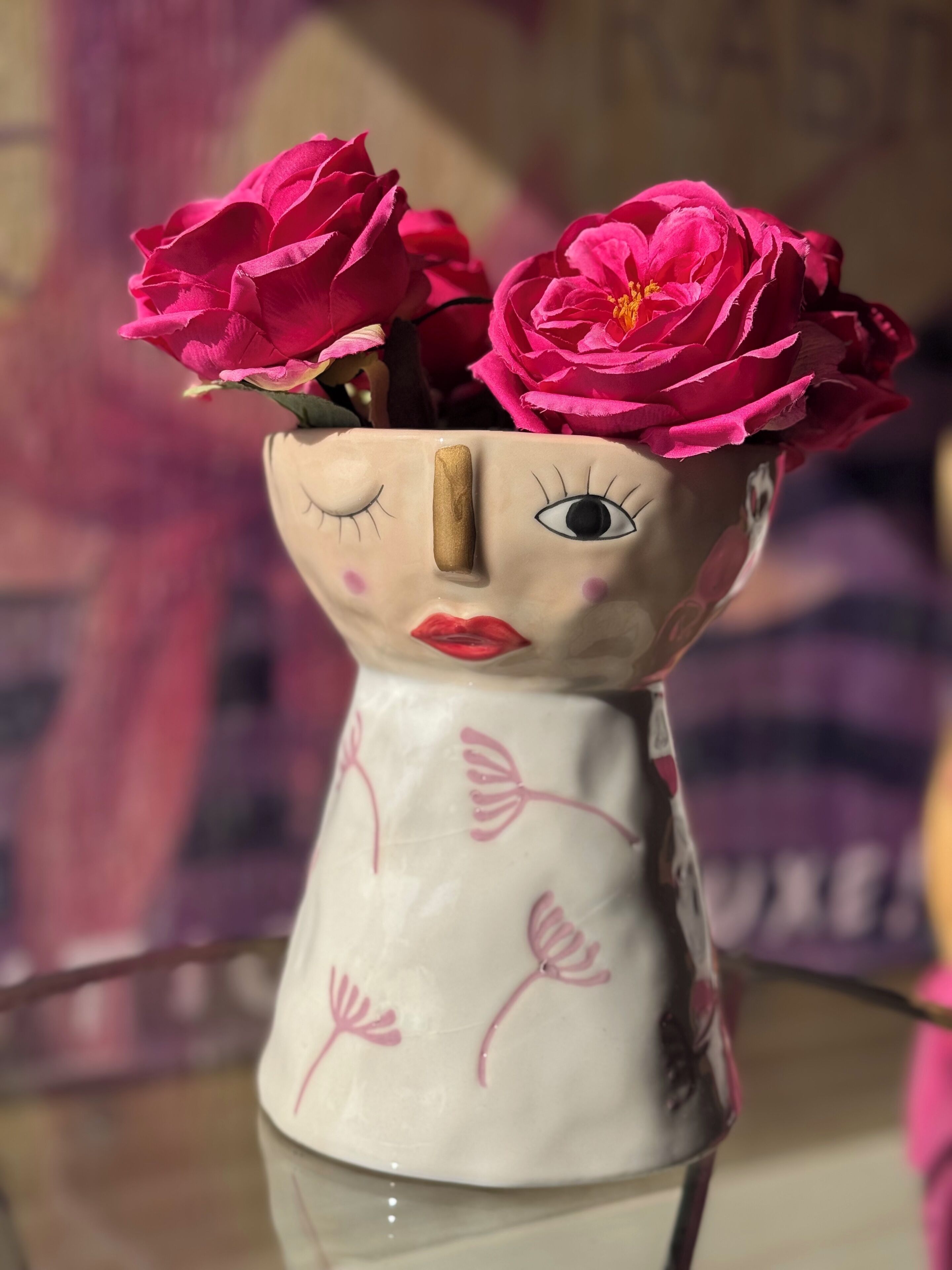 Un jarrón de cerámica con diseño facial, que contiene flores rosas.