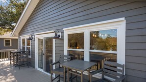 Un patio techado al aire libre con mesas y sillas de madera, una entrada por puerta de vidrio y una ventana con lámpara.