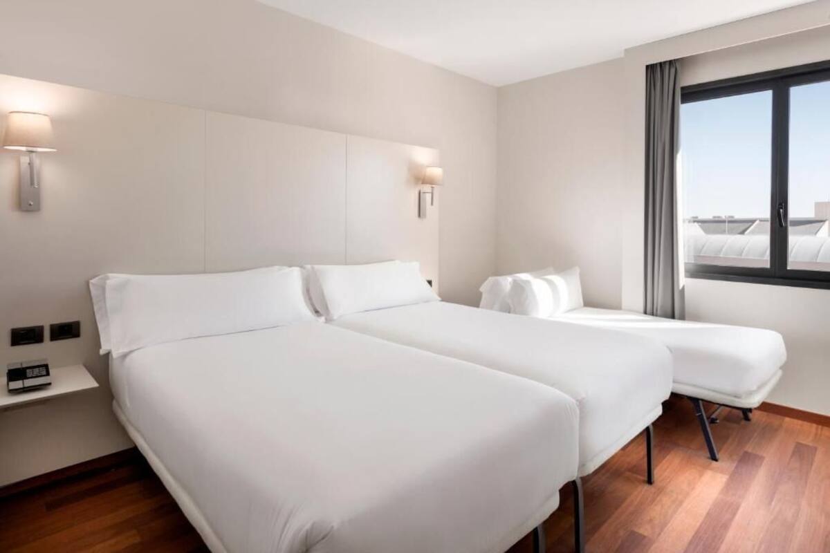 Habitación de hotel con dos camas, una mesita de noche con lámpara, ventana con vistas y suelo de madera.