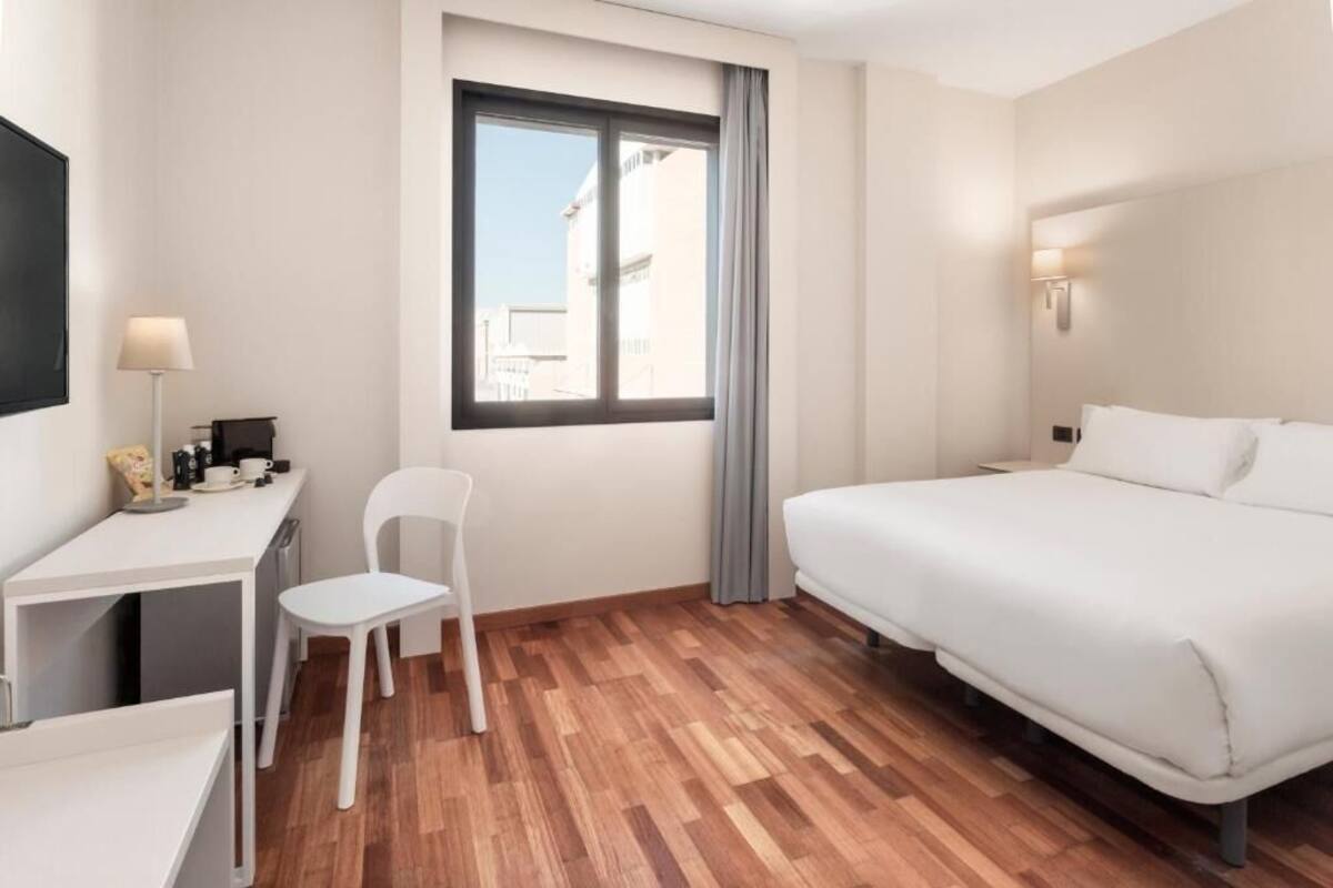 Habitación de hotel con una cama, un escritorio con silla, una lámpara y una ventana con cortinas.