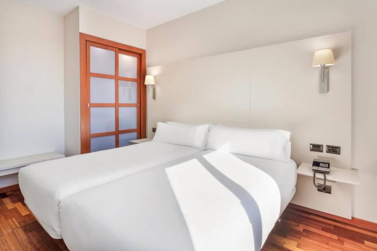 Una habitación de hotel con una cama grande, una puerta de madera, una mesita de noche con lámpara y un teléfono fijo en la pared.