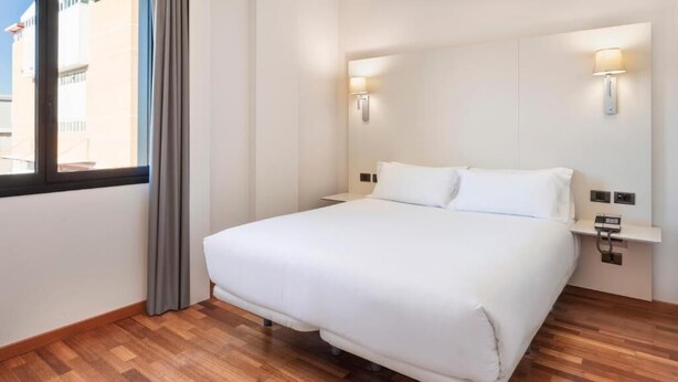 A hotel room with a large bed, wooden flooring, two wall-mounted light fixtures, and a window with curtains.