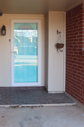 A front door with a glass panel, a small porch with steps, a wall-mounted lantern, and a brick wall.