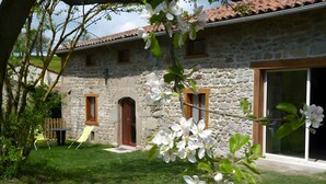 Une maison en pierre avec une porte rouge, un petit patio avec des chaises et un jardin parsemé de fleurs blanches.