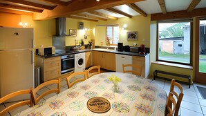 Une cuisine avec des poutres en bois, une table à manger recouverte d’une nappe à motifs floraux et une fenêtre donnant sur l’extérieur.