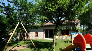 Un jardin avec une balançoire, un toboggan, une petite maison en pierre et un petit chalet de jeux.