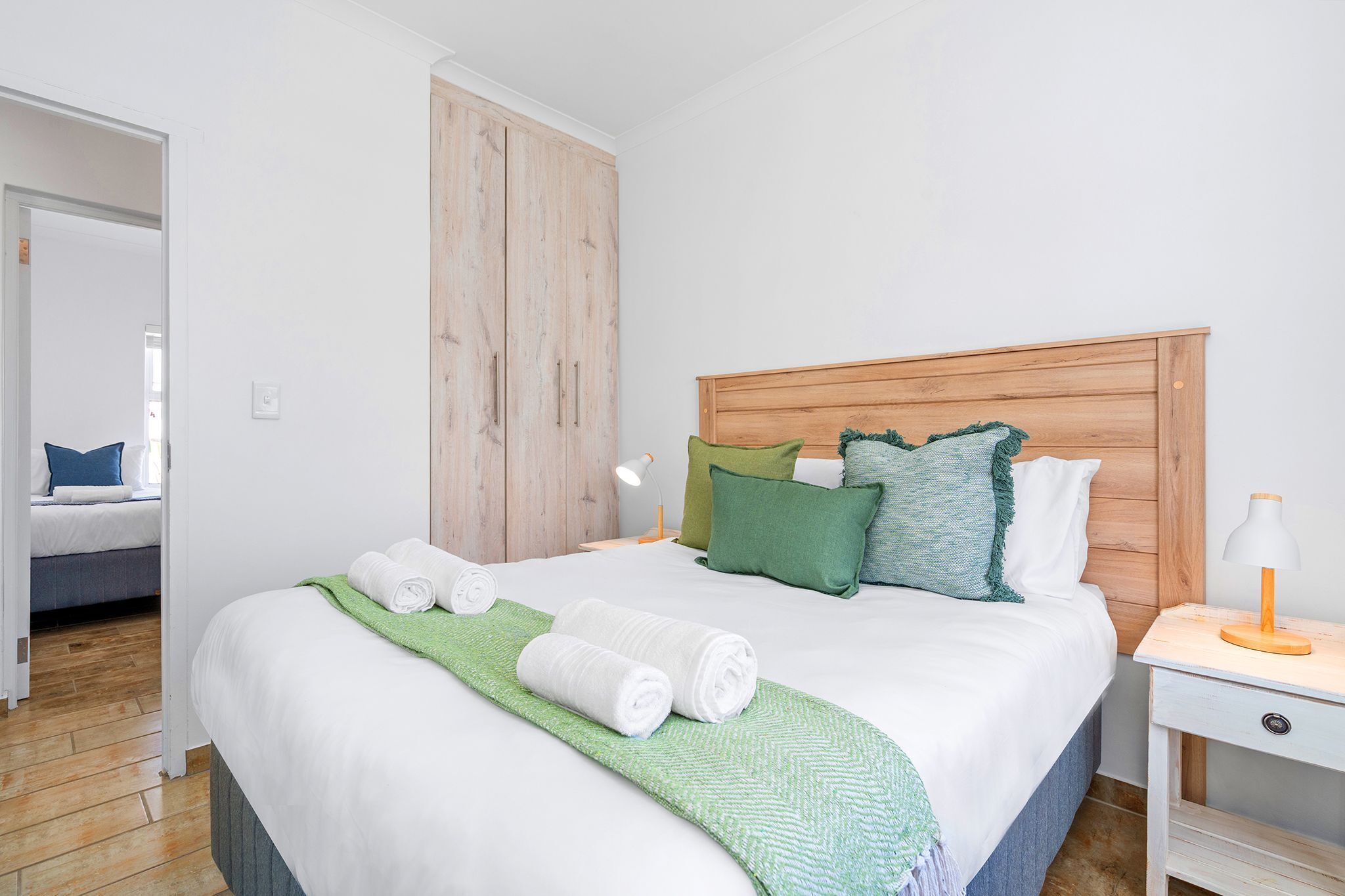 A hotel room with a wooden headboard, a bed with white bedding, green and white pillows, a nightstand with a lamp, and a green towel on the bed.