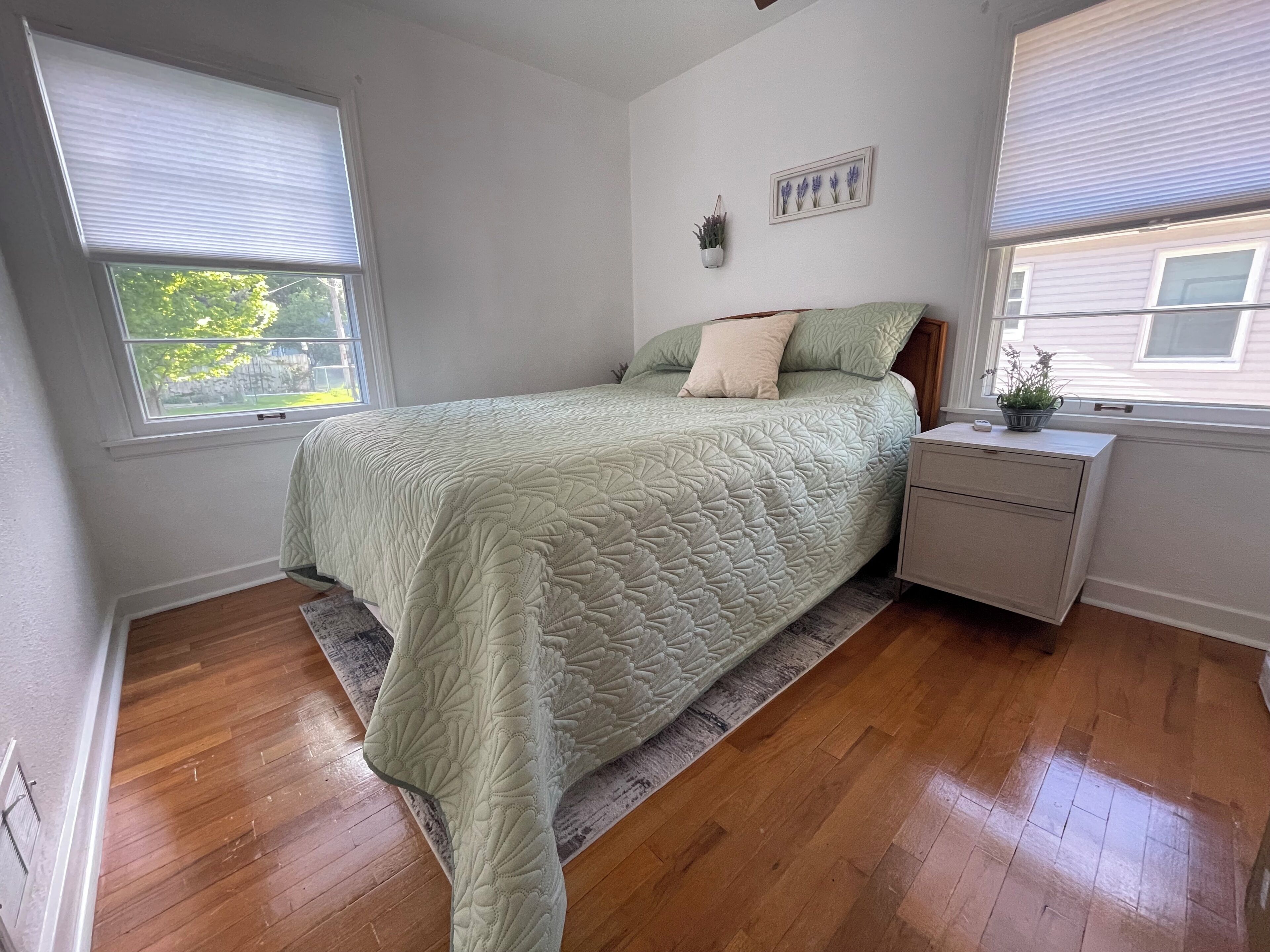 Une chambre à coucher comprenant un lit, une table de chevet, une fenêtre avec des stores, une plante fixée au mur et un tableau encadré.