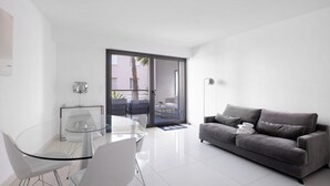 Un salon moderne avec une table à manger en verre, des chaises blanches, un canapé gris et une porte-fenêtre coulissante donnant sur un espace extérieur.