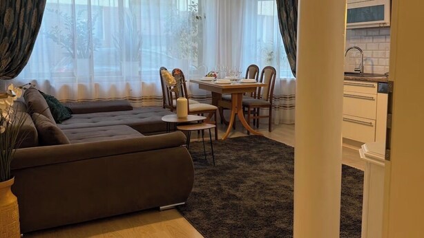 Ein modernes Wohnzimmer mit einer Couch, einem Esstisch und Stühlen, mit einer durch eine offene Tür sichtbaren Küche und großen Fenstern mit durchsichtigen Vorhängen.